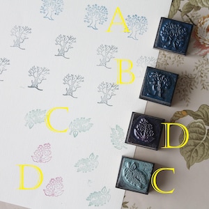 Puede incluir: Un surtido de cuatro sellos de tinta cuadrados de color azul oscuro con diseños de árboles y hojas. Los sellos están junto a múltiples impresiones de árboles y hojas en tinta azul, verde y morada sobre papel blanco. El fondo presenta papel pintado floral.