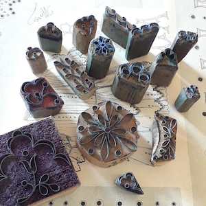 Può includere: Una collezione di timbri vintage in legno e metallo con vari disegni floreali e geometrici. I timbri sono di varie forme e dimensioni, con blocchi di legno scuro e intricati motivi metallici. Alcuni timbri hanno una tonalità viola o rossa.