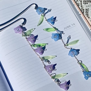 May include: Two metal bookmarks with floral designs. One bookmark features purple flowers, while the other has blue flowers. Both have green leaf accents and are on a notebook page with the dates "JUNE 13" and "JUNE 14".