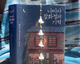 May include: A book titled "Namiya General Store's Miracle" with Korean text and a decorative bookmark. The cover depicts a building with a red bicycle and a cat. The bookmark has white flower charms.