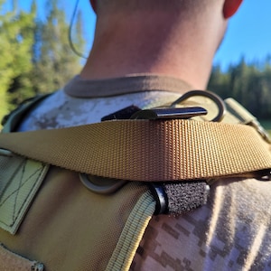 May include: Close-up of a tan tactical vest with a shoulder strap. The strap is made of a woven material and features a black plastic buckle and a metal loop. The vest is worn over a camouflage shirt, with a green field and blue sky in the background.