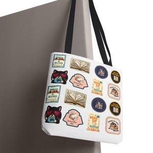 May include: White tote bag with black handles featuring various book-themed graphics. Designs include a cat wearing heart-shaped sunglasses reading a book, open books, and text like "Booked & Blessed" and "Read, Sip, Repeat."