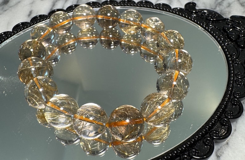 May include: A clear quartz bracelet with golden rutile inclusions, displayed on a reflective surface. The bracelet features large, round beads strung together with a golden elastic cord. The beads are approximately 1.5 cm in diameter. The bracelet is set against a black, ornate frame.