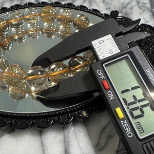 May include: A digital caliper measures a bracelet of clear, round beads with golden inclusions. The bracelet is reflected in a round, black-framed mirror. The caliper displays a measurement of 13.5 mm.