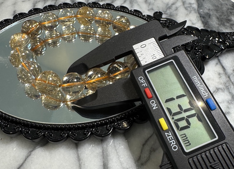 May include: A digital caliper measures a bracelet of clear, round beads with golden inclusions. The bracelet is reflected in a round, black-framed mirror. The caliper displays a measurement of 13.5 mm.