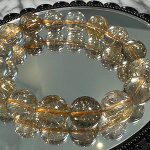 May include: A clear quartz bracelet with golden rutile inclusions, resting on a reflective surface within a black, ornate frame. The bracelet's beads are spherical and reflect light, creating a shimmering effect.