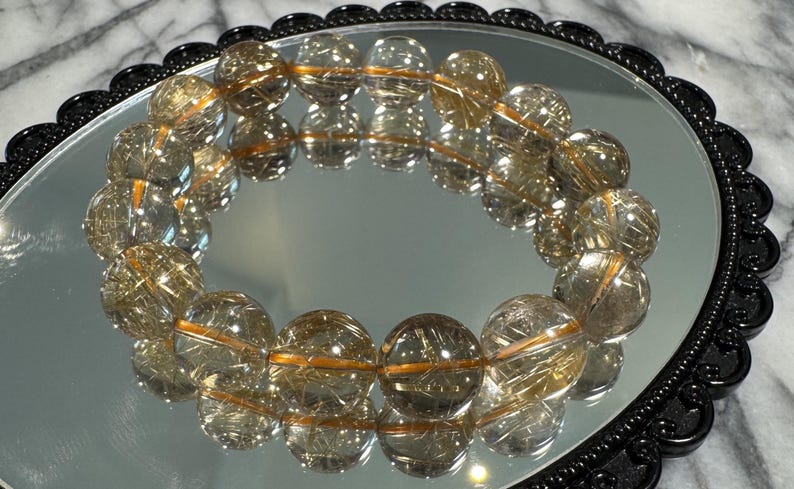 May include: A clear quartz bracelet with golden rutile inclusions, resting on a reflective surface within a black, ornate frame. The bracelet's beads are spherical and reflect light, creating a shimmering effect.