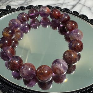 Peut inclure: Un bracelet de pierres précieuses rondes et polies dans les tons violets et bruns, se reflétant sur une surface miroitante. Les perles sont disposées en cercle. Le bracelet est présenté sur un miroir noir ovale orné.