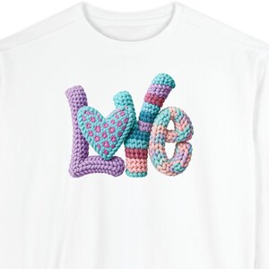 May include: White long-sleeved sweatshirt featuring the word "Love" in colourful, textured, crochet letters. The "L" is purple, the heart is blue with a leopard print, the "o" is striped, and the "e" is multi-coloured.