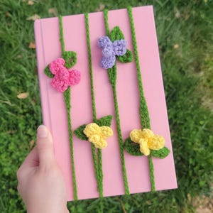 May include: A pink notebook decorated with crocheted flowers in pink, purple, and yellow. Green yarn stems connect the flowers. The notebook is held against a background of green grass.