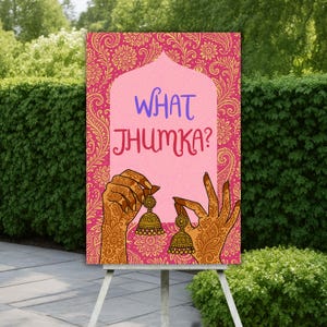 Op de afbeelding: Een roze bord met de tekst "WHAT JHUMKA?" en illustraties van handen die gouden oorbellen vasthouden. Het bord heeft een bloemenpatroon en wordt buiten op een wit schildersezel tentoongesteld.