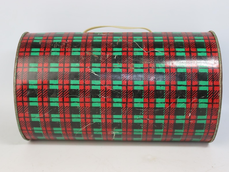 May include: A cylindrical tin with a red, black, and green plaid pattern. The tin has a beige handle and a metallic rim. The plaid design is a classic pattern, and the tin appears to be vintage.