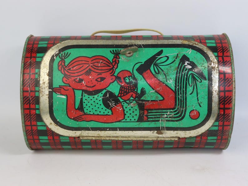 May include: Vintage metal lunchbox with a green, red, and black plaid pattern. The front features a cartoon illustration of a girl with red hair and a small creature. The lunchbox has a yellow handle and a metal clasp.
