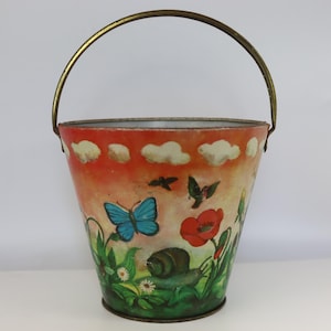May include: A metal bucket featuring a painted scene of a bright blue butterfly, a snail, a red poppy, and a hummingbird against an orange sky with white clouds. The bucket has a thin, curved brass-colored metal handle attached to the top rim.
