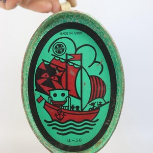 May include: Vintage oval tin with a green border and a central illustration of a red sailboat with black and red sails. The tin has the text "MADE IN USSR" and a logo. The bottom of the tin has the text "U.-50".