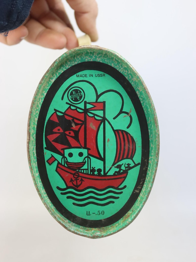 May include: Vintage oval tin with a green border and a central illustration of a red sailboat with black and red sails. The tin has the text "MADE IN USSR" and a logo. The bottom of the tin has the text "U.-50".