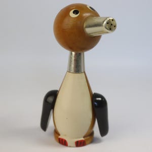 May include: A vintage penguin-shaped salt or pepper shaker. The shaker features a wooden head and base, a cream-colored body, and black wings. The beak is a silver-colored metal with holes for dispensing.