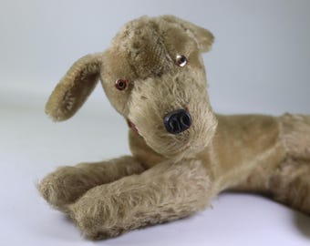 May include: A vintage plush dog toy in a lying position. The dog has tan fur, a black nose, and red eyes. It has floppy ears and a red collar. The toy is made of a soft, fuzzy material.