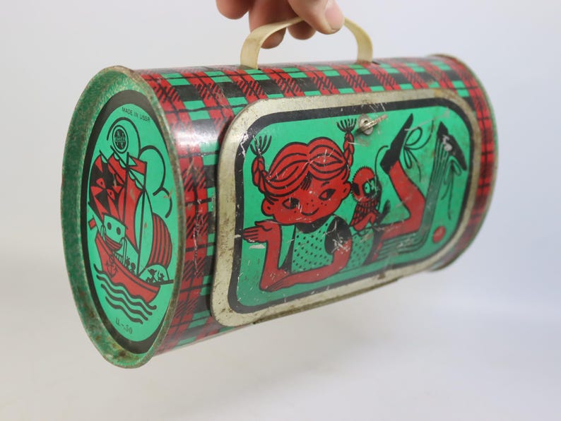 May include: Vintage metal lunchbox with a red and green plaid pattern. The ends feature a green background with a red sailboat illustration. The front has a cartoon girl and a monkey. Includes a white handle.