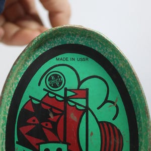 May include: Vintage green and black metal tray with a red and black sailboat design. The tray has the text "MADE IN USSR" and the "NORMA" logo. The tray is oval-shaped and has a worn, vintage appearance.