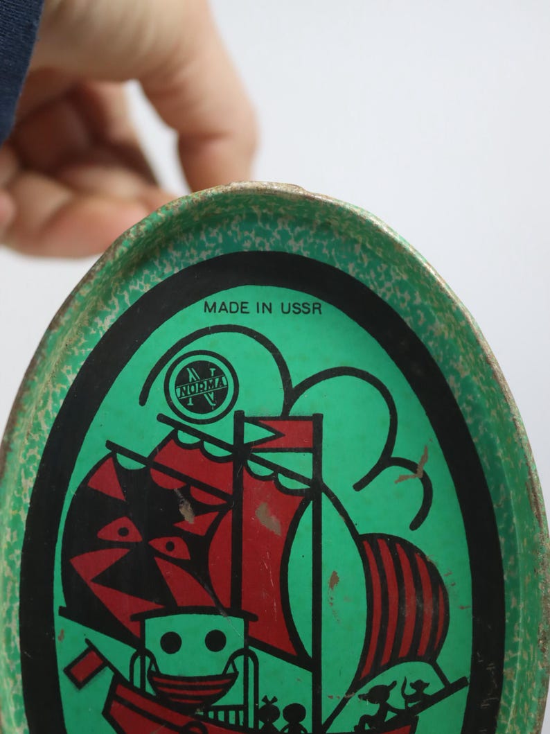 May include: Vintage green and black metal tray with a red and black sailboat design. The tray has the text "MADE IN USSR" and the "NORMA" logo. The tray is oval-shaped and has a worn, vintage appearance.