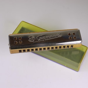 May include: A silver-toned harmonica with the word "Hohner" engraved on the top, resting on a translucent yellow-green case. The harmonica has a wooden base and multiple holes for sound production. Musical instrument.