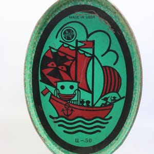 May include: Vintage oval tin with a green background and a red and black sailboat illustration. The tin has the text "MADE IN USSR" and "Ц.-50". The illustration features a ship with red sails and a red anchor.