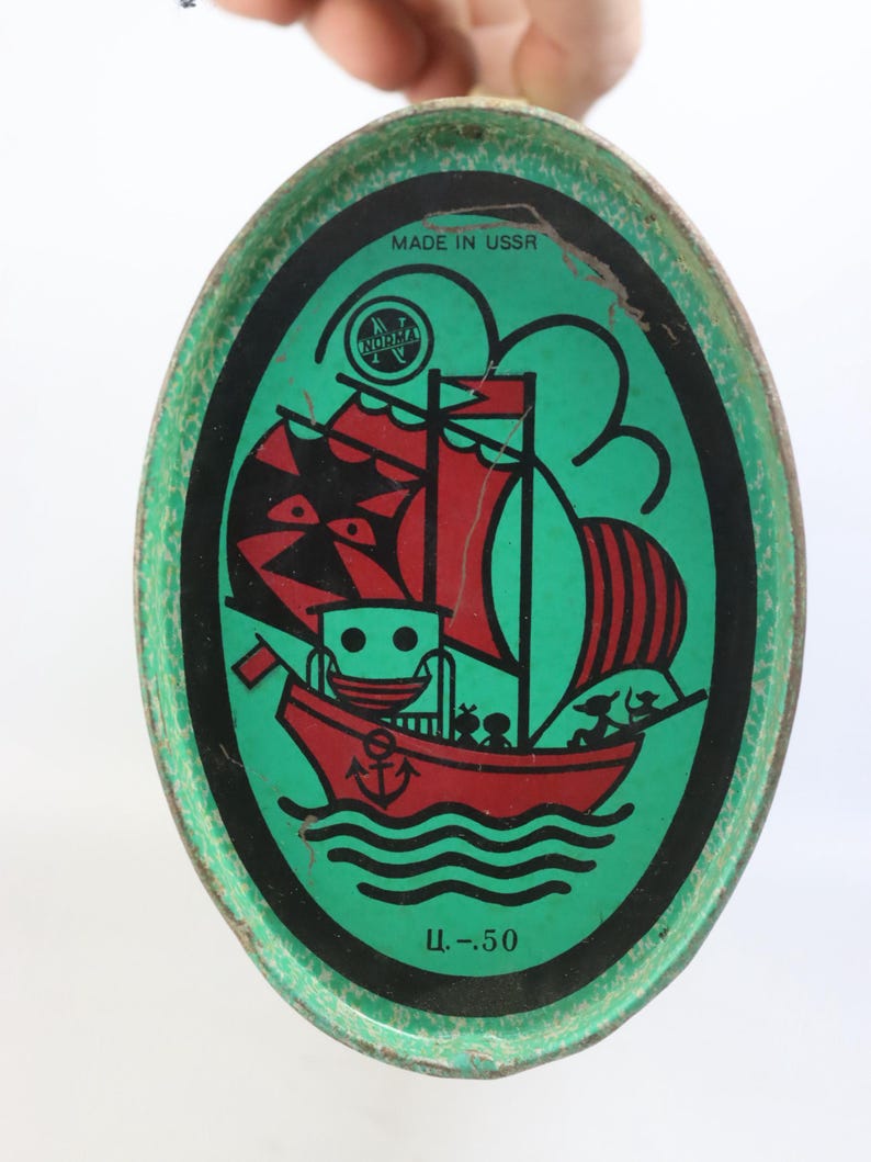 May include: Vintage oval tin with a green background and a red and black sailboat illustration. The tin has the text "MADE IN USSR" and "Ц.-50". The illustration features a ship with red sails and a red anchor.