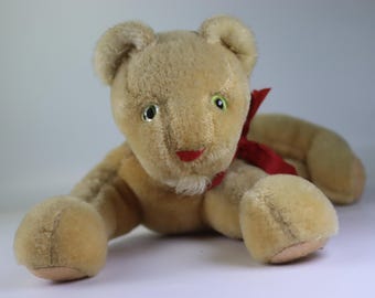 Könnte beinhalten: Ein Vintage-Plüschlöwenspielzeug in liegender Position. Der Löwe hat hellbraunes Fell, grüne Augen und eine rote Schleife um den Hals. Das Spielzeug hat einen roten Mund und einen kleinen weißen Bart.