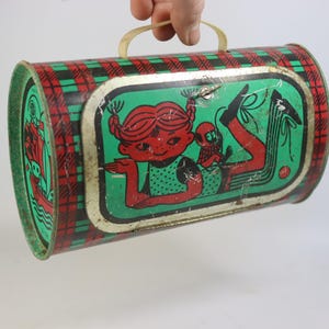 May include: Vintage metal lunchbox with a red and green plaid pattern on the sides. The front features a cartoon girl with red hair and a monkey on a green background. It has a yellow plastic handle.