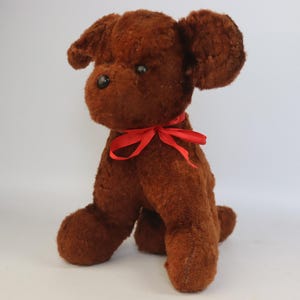 May include: A brown plush dog toy with a red ribbon tied around its neck. The dog has floppy ears and black button eyes. The toy is sitting upright, with its front paws forward.