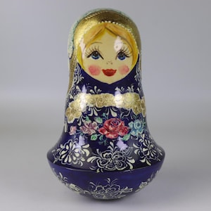 May include: A hand-painted Russian nesting doll. The doll has a light-skinned face with blue eyes, red lips, and rosy cheeks. The head is gold, and the body is navy blue with floral and gold leaf accents. A traditional folk art item.