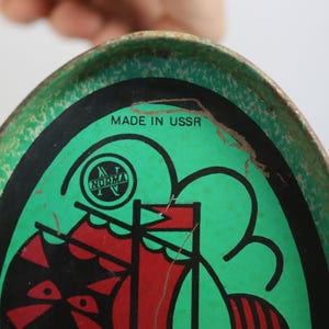 May include: Vintage oval metal tray with a green background and a black and red graphic of a ship. The tray has the text "MADE IN USSR" and a logo that says "NORMA". The tray has a distressed look.