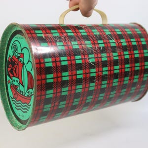 May include: Vintage cylindrical tin container with a red, black, and green plaid pattern. The ends are green with a nautical illustration of a ship. It has a cream-colored handle. Made in USA.