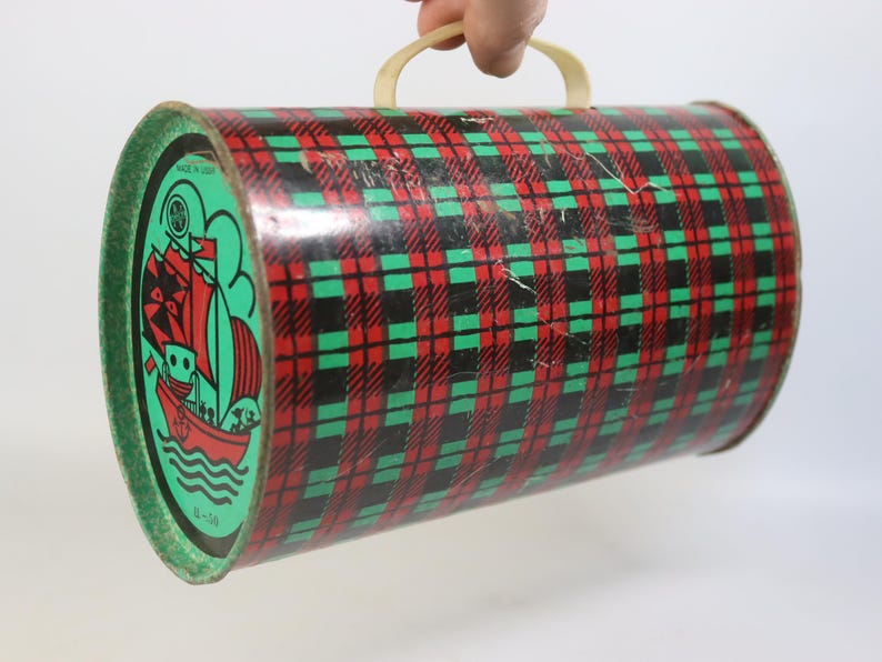 May include: Vintage cylindrical tin container with a red, black, and green plaid pattern. The ends are green with a nautical illustration of a ship. It has a cream-colored handle. Made in USA.
