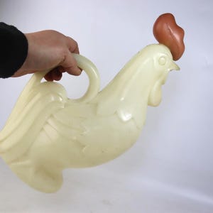 May include: A vintage, off-white plastic rooster-shaped pitcher with a brown comb. The pitcher has a handle and is held by a person. The rooster has detailed feather and facial features.