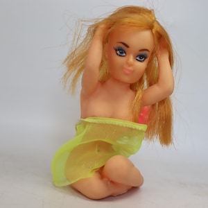 May include: A vintage doll with blonde hair and blue eyes, wearing a yellow mesh skirt. The doll is kneeling with its hands in its hair, and has a pink accent on its chest. The doll is on a white background.