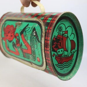 May include: Vintage metal lunchbox with a red, green, and black plaid pattern. The lunchbox features illustrations of a mermaid and a ship with a smiling face. The handle is cream-colored, and the lunchbox is marked "MADE IN USA".