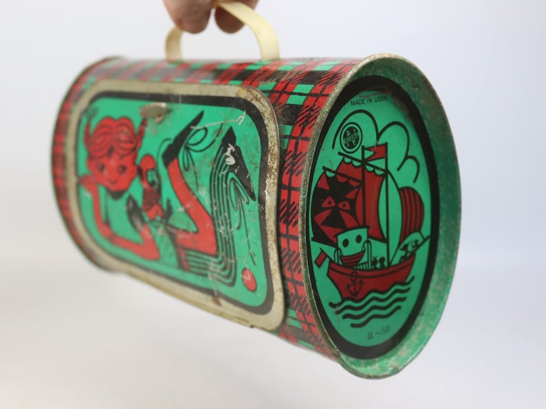 May include: Vintage metal lunchbox with a red, green, and black plaid pattern. The lunchbox features illustrations of a mermaid and a ship with a smiling face. The handle is cream-colored, and the lunchbox is marked "MADE IN USA".