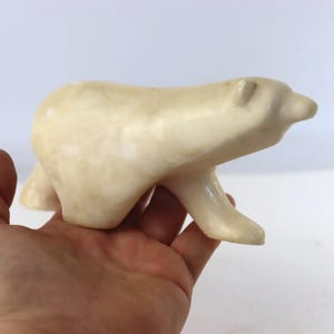 May include: A smooth, off-white stone carving of a polar bear in a walking position. The sculpture features a polished, marbled surface with subtle variations in tone, capturing the animal's rounded form, small ears, and extended snout in detail.