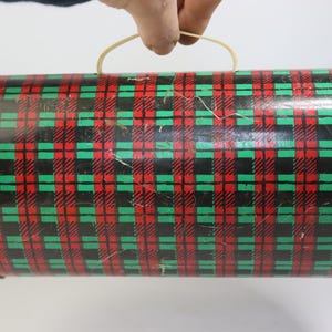 May include: A cylindrical tin container with a red, green, and black plaid pattern. The container has a curved handle made of a light-colored material. The tin appears to be vintage or antique.