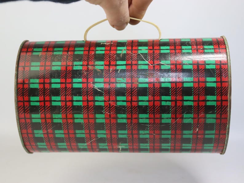 May include: A cylindrical tin container with a red, green, and black plaid pattern. The container has a curved handle made of a light-colored material. The tin appears to be vintage or antique.