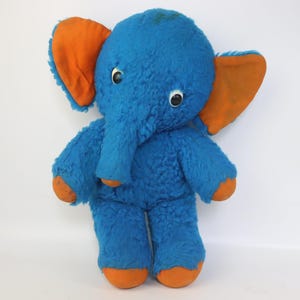Op de afbeelding: Een vintage blauwe pluche olifant met oranje oren, voeten en slurf. Het knuffeldier heeft grote, ronde ogen en een zachte, fuzzy textuur. Een klassiek kinderspeelgoed.