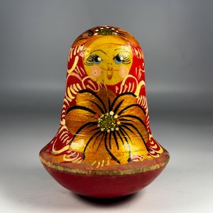 Könnte beinhalten: Handbemalte russische Babuschka-Puppe in Rot, Gelb und Gold. Die Puppe hat ein lächelndes Gesicht mit blauen Augen und ein großes Blumendesign. Der Sockel ist rot mit goldenem Rand, typisch für dieses Handwerk.