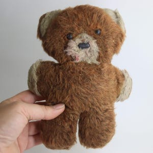 May include: A vintage brown teddy bear with tan accents on the ears, paws, and snout. The plush toy has a worn appearance, with visible wear and tear on the fabric. The bear has black button eyes and a black nose.