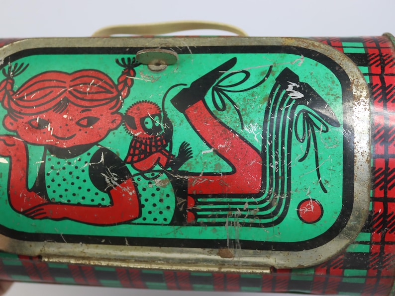 May include: Vintage metal lunchbox with a green and red plaid pattern. The lid features a cartoon girl with red hair and a monkey, set against a green background. The lunchbox has a metal handle and a latch closure.
