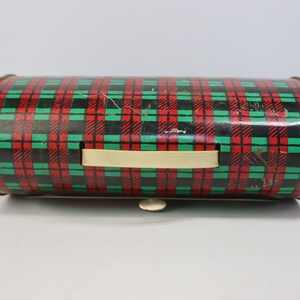 May include: A cylindrical metal container with a red, green, and black plaid pattern. It has a cream-colored handle and a small, silver-colored latch. The container appears to be vintage or antique.