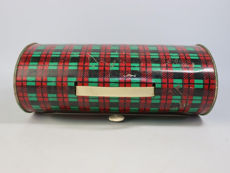 May include: A cylindrical metal container with a red, green, and black plaid pattern. It has a cream-colored handle and a small, silver-colored latch. The container appears to be vintage or antique.