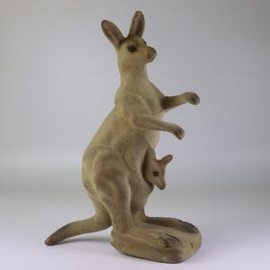 May include: A vintage kangaroo figurine in a standing pose, crafted from a light brown material. The kangaroo has a joey in its pouch. The figurine has a textured surface and is set against a plain background.