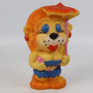 Puede incluir: Figura de león vintage de estilo caricatura. El león tiene pelo naranja, ojos azules y labios rojos. Sostiene una sombrilla rosa y lleva pantalones cortos azules. La figura es principalmente amarilla y naranja, con detalles rojos y azules.